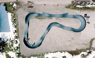 Eine Draufsicht auf eine geschwungene Rennbahn, die aus schwarzem Material besteht. Rund um die Bahn sind einige Menschen und Schneereste zu sehen. | © Christian Posch | Klaus Zopf