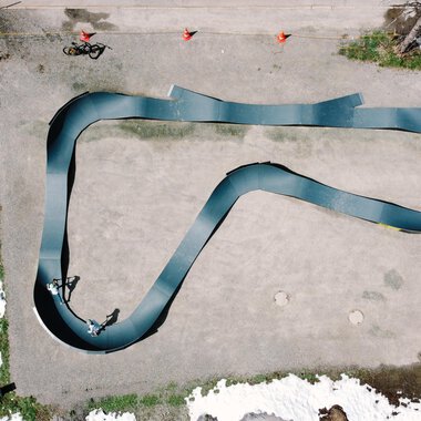 A top view of a curved racetrack made of black material. Around the track, some people and remnants of snow can be seen. | © Christian Posch | Klaus Zopf