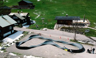 Eine moderne, geschwungene Rodelbahn in einer malerischen Berglandschaft. Im Hintergrund sind rustikale Holzhäuser und grüne Wiesen zu sehen. | © Christian Posch | Klaus Zopf