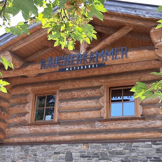 Ein rustikales Holzgebäude mit einem Schild, das "Mühlenkammer" zeigt. Umgeben von grünen Blättern und einem klaren Himmel. | © Kleinwalsertal Tourismus | Veronika Senn