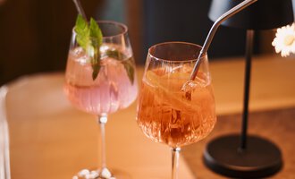 Two elegant cocktails are on a wooden table. One is pink with mint, the other is orange and has a straw.