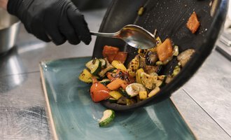 A cook serves roasted vegetables from a pan onto a plate. The colorful vegetable pieces are attractively arranged.
