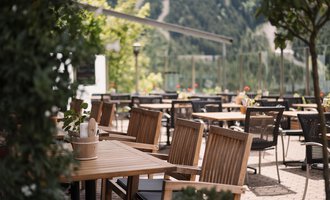 A cozy outdoor area with wooden tables and chairs. In the background are green trees and a mountain landscape.
