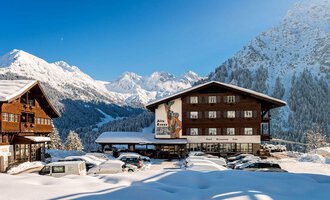 A charming Alpine landscape in winter with snow-covered mountains. In the foreground, there is a hotel and several parked cars.