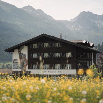 Ein traditionelles Gebäude in einer malerischen Landschaft. Im Vordergrund blühen gelbe Wildblumen, während die Berge im Hintergrund sichtbar sind.