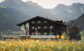 Ein traditionelles Gebäude in einer malerischen Landschaft. Im Vordergrund blühen gelbe Wildblumen, während die Berge im Hintergrund sichtbar sind.