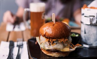 Ein saftiger Burger mit geschmolzenem Käse und frischem Gemüse auf einem Holzbrett. Im Hintergrund ist ein Glas Bier zu sehen.