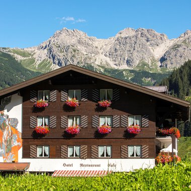Ein charmantes Chalet in den Bergen mit Blumenbalkonen. Im Hintergrund sind beeindruckende Berggipfel und ein klarer Himmel zu sehen. | © Hotel Alte Krone