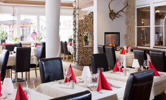 An elegant restaurant with modern tables and chairs. The tablecloths are cream-colored and the napkins are in red, while a fireplace in the background creates a cozy atmosphere. | © Aparthotel Kleinwalsertal Haller GmbH & Co KG