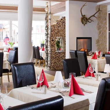 Ein elegantes Restaurant mit modernen Tischen und Stühlen. Die Tischdecken sind cremefarben und die Servietten in Rot, während ein Kamin im Hintergrund für eine gemütliche Atmosphäre sorgt. | © Aparthotel Kleinwalsertal Haller GmbH & Co KG