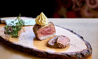 A piece of tender meat on a wooden board, garnished with mustard and fresh herbs. In the background, a stylishly patterned textile can be seen. | © Hotel Tradizio | © Annette Sandner