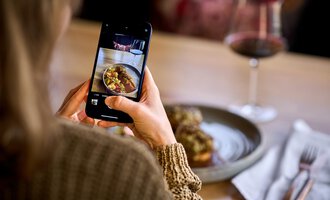 A person is taking a photo of their food with a smartphone. In the background, a glass of wine and a set table can be seen. | © Hotel Tradizio | © Annette Sandner