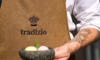 Ein Kellner hält eine Schale mit drei verschiedenen Eissorten. Er trägt eine braune Schürze mit dem Logo "tradizio". | © Hotel Tradizio | © Annette Sandner