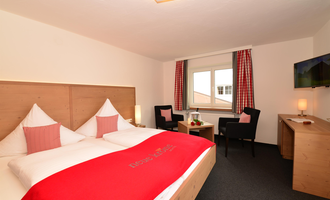 Ein gemütliches Hotelzimmer mit einem Doppelbett, zwei Stühlen und einem Tisch. Die Wände sind hell, und der Raum hat große Fenster mit Vorhängen.