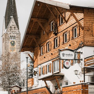 A traditional wooden house in the snow with a bell tower in the background. The scene conveys a cozy winter atmosphere.