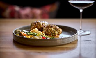 An elegantly arranged plate with two crispy delicacies and colorful vegetables. In the background, there is a glass of wine. | © Hotel Tradizio | © Annette Sandner