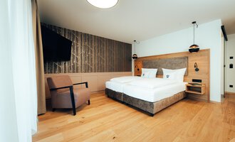 A modern hotel room with a large double bed and a cozy armchair. The walls are painted in neutral tones, creating a relaxed atmosphere.