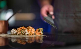 A cook is preparing fresh shrimp on a grill surface. The background is blurred, which draws the focus to the prepared dishes.