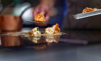 A hand serves grilled shrimp on a hot surface. In the background, a pot can be seen.