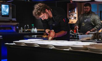 A cook is preparing dishes in a modern kitchen. In the background, another cook is watching.