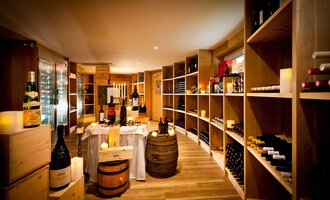 Ein gemütlicher Weinkeller mit Holzregalen voller Weinflaschen. In der Mitte steht ein Tisch mit Kerzen und weiteren Weinen. | © Hotel Birkenhöhe | Fotograf Krug