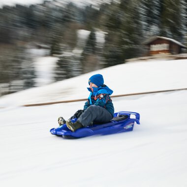 Kinder ziehen Schlitten im Schnee, während Erwachsene in warmen Kleidern sie begleiten. Im Hintergrund ist eine Kirche und der blaue Himmel zu sehen. | © Kleinwalsertal Tourismus | Oliver Farys