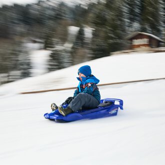 Kinder ziehen Schlitten im Schnee, während Erwachsene in warmen Kleidern sie begleiten. Im Hintergrund ist eine Kirche und der blaue Himmel zu sehen. | © Kleinwalsertal Tourismus | Oliver Farys