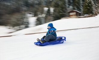 Kinder ziehen Schlitten im Schnee, während Erwachsene in warmen Kleidern sie begleiten. Im Hintergrund ist eine Kirche und der blaue Himmel zu sehen. | © Kleinwalsertal Tourismus | Oliver Farys