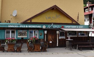 Ein gemütliches Gasthaus mit einem bunten Schild und blühenden Blumen auf der Terrasse. Die Fassade ist in warmen Farben gehalten und vermittelt eine einladende Atmosphäre. | © s‘Älpele | Marco Mitterer