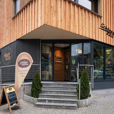 Ein modernes Restaurant mit Holzfassade und großen Fenstern. Der Eingangsbereich ist einladend gestaltet, mit einem Schild für das Restaurant. | © Schönegger Käse-Alm