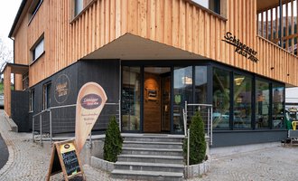 Ein modernes Gebäude mit Holzfassade und großen Fenstern. Vor dem Eingang steht ein Schild mit einer Einladung. | © Schönegger Käse-Alm