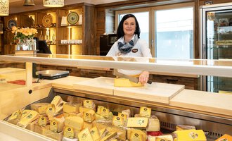 Eine Käseverkäuferin steht hinter einem Verkaufstresen mit einer großen Auswahl an Käse. Der Laden hat eine helle und einladende Atmosphäre. | © Schönegger Käse-Alm