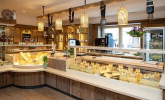 A cheese shop with various types of cheese on wooden shelves. The decor is rustic and inviting, with warm lighting and decorative elements. | © Schönegger Käse-Alm