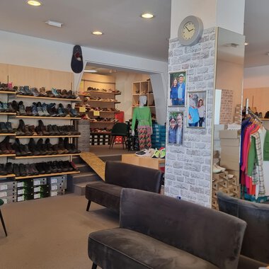 Ein Schuhgeschäft mit zahlreichen Regalen voller Schuhe. In der Mitte stehen bequeme Sofas und Kleidung ist zur Schau gestellt. | © Schuhhaus Schuster | Marietta Diehl