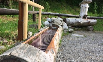 Ein rustikaler Holzbrunnen aus Baumstämmen in einer natürlichen Umgebung. Im Hintergrund sind weitere Holzelemente und Vegetation zu sehen. | © Kleinwalsertal Tourismus