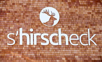 Ein stilvolles Schild mit dem Schriftzug "s'hirscheck". Der Hintergrund besteht aus Holzschindeln und zeigt ein Hirsch-Logo. | © Kleinwalsertal Tourismus | Frank Drechsel