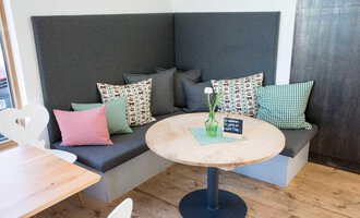 Ein gemütlicher Cafébereich mit einer gepolsterten Bank und bunten Kissen. Auf dem Tisch steht eine kleine Vase mit einer Blume. | © Kleinwalsertal Tourismus | Frank Drechsel