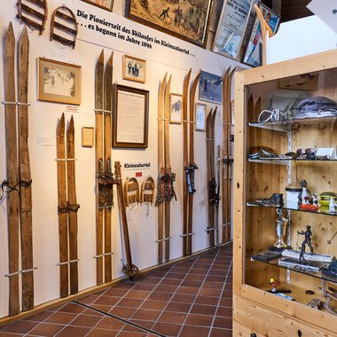 Eine Ausstellung mit mehreren Holzskiern, die an Wänden ausgestellt sind. Im Hintergrund sind Fotos und Informationen über die Skigeschichte sichtbar. | © Kleinwalsertal Tourismus | Oliver Farys