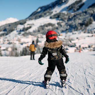 Two skiers walk along a snow-covered path. In the background, trees and a clear blue sky are visible. | © Kleinwalsertal Tourismus | Oliver Farys