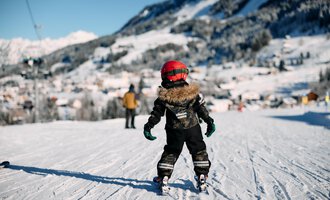 Two skiers walk along a snow-covered path. In the background, trees and a clear blue sky are visible. | © Kleinwalsertal Tourismus | Oliver Farys