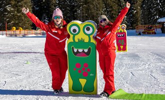 Two cheerful people in red suits are standing next to a colorful monster sign in the snow. In the background, trees and a snowy landscape can be seen. | © Frank Drechsel alpin.photo