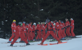 A group of skiers in red suits stands in a snowy ski area. The trees in the background create a winter atmosphere. | © Skischule Hirschegg GmbH