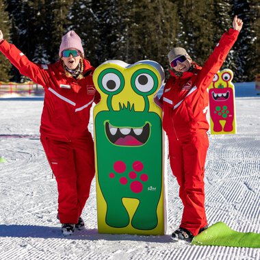 Zwei fröhliche Menschen in roten Anzügen stehen neben einem bunten Monster-Schild im Schnee. Im Hintergrund sind Bäume und eine verschneite Landschaft zu sehen. | © Frank Drechsel alpin.photo