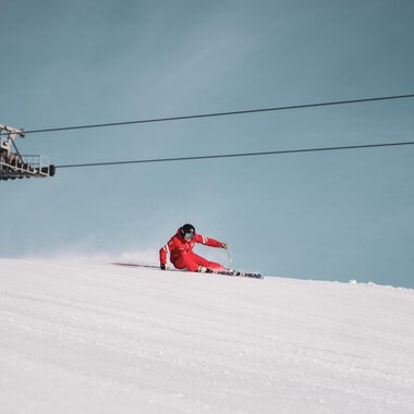 A skier in a red suit is skiing down the slope. In the background, a chairlift can be seen against a clear blue sky.