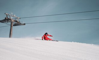 A skier in a red suit is skiing down the slope. In the background, a chairlift can be seen against a clear blue sky.