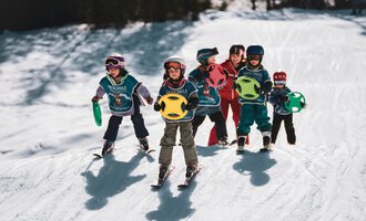 Six children in ski gear are standing on a snow hill holding colorful play balls. The sun is shining and the surroundings are covered in snow.
