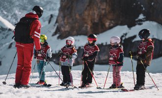 A group of children is standing on skis in the snow while being instructed by a ski teacher. In the background, mountains and a clear sky can be seen.