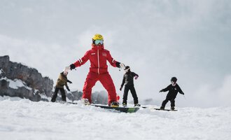 A ski instructor in red clothing is standing on a snowboard. Behind him, three children are practicing on the slope.