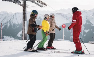 A group of skiers is standing on a snow-covered slope. In the background, there are mountains and a ski lift.