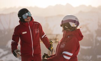 Two skiers in red jackets are happily chatting in a snowy mountain landscape. In the background, snow-covered mountains and a clear sky can be seen.